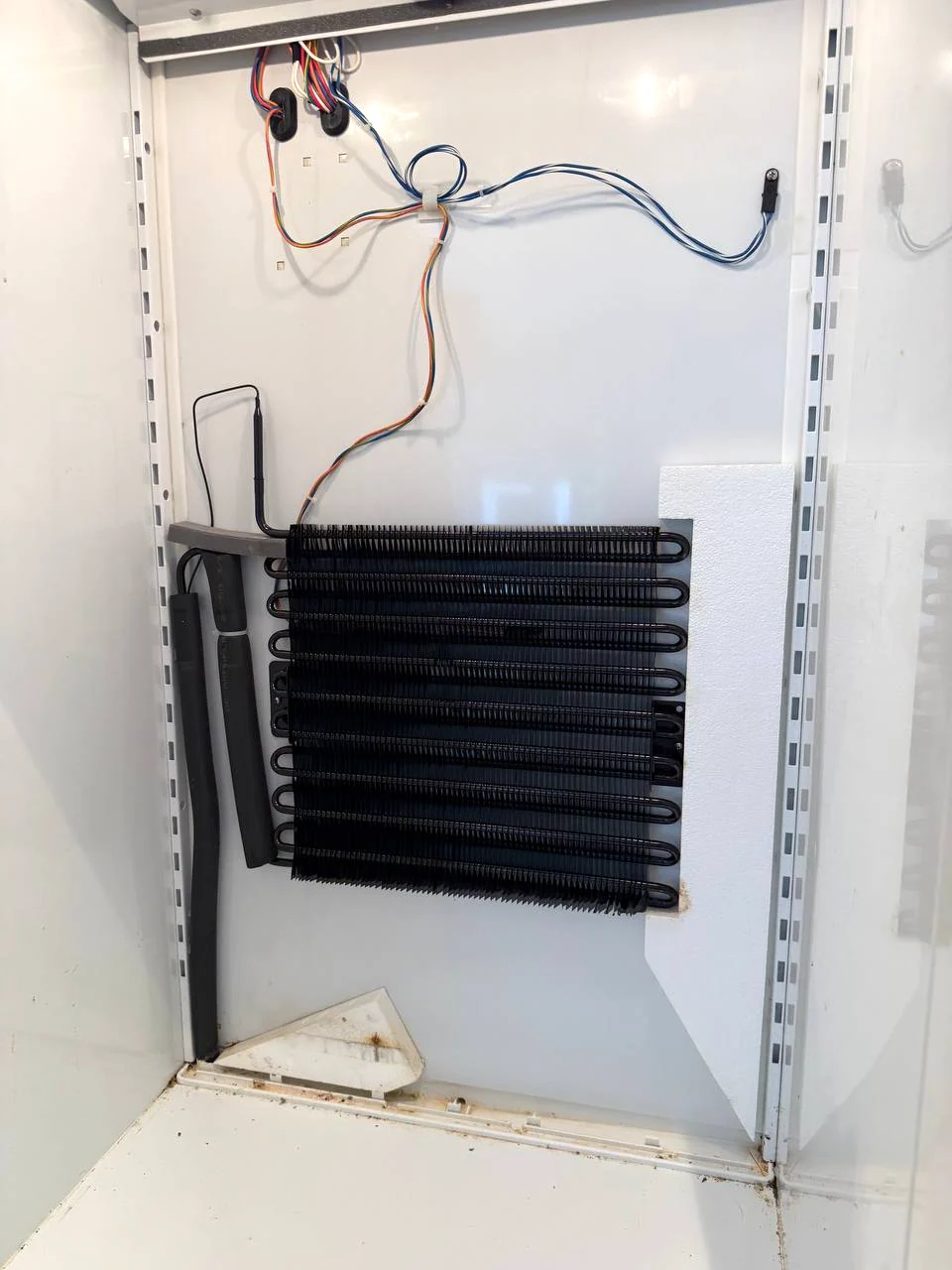 Black evaporator coil on the rear wall of a refrigerated cabinet with loose sensor wires