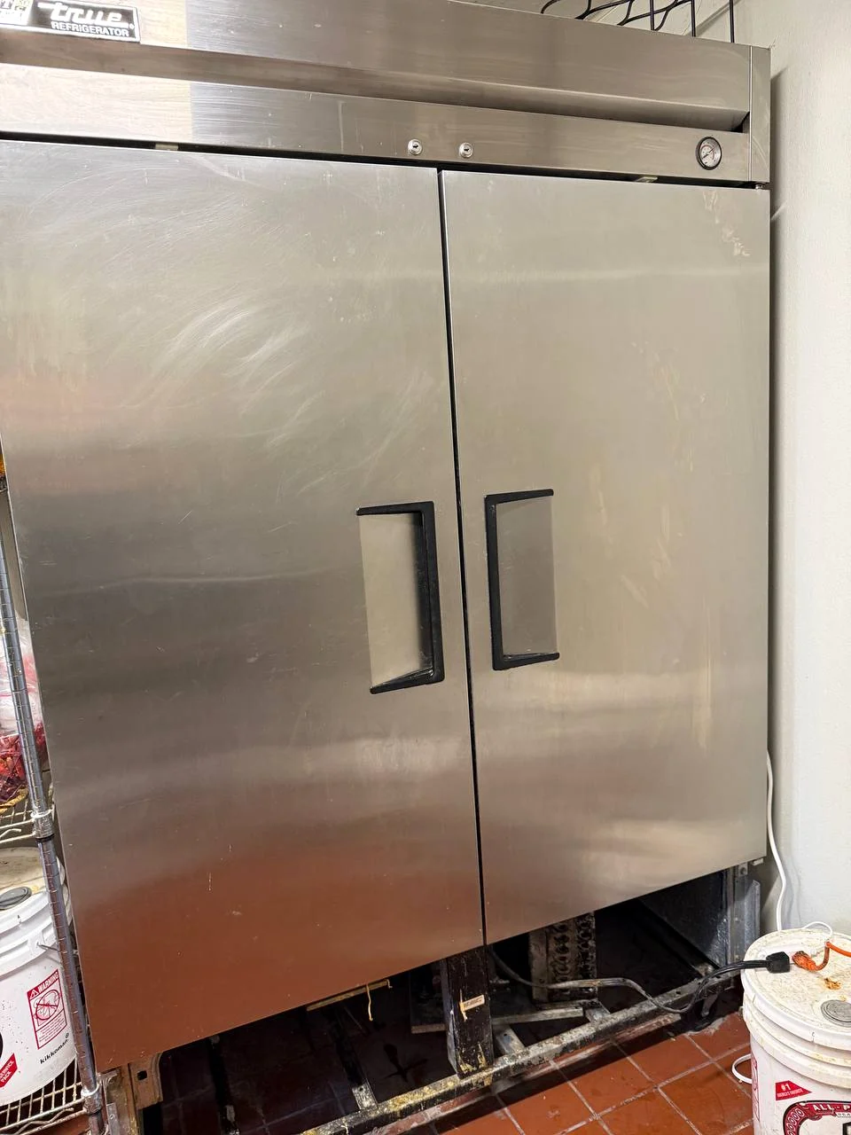 Two-door stainless reach-in refrigerator with the lower panel off and a dirty compressor area